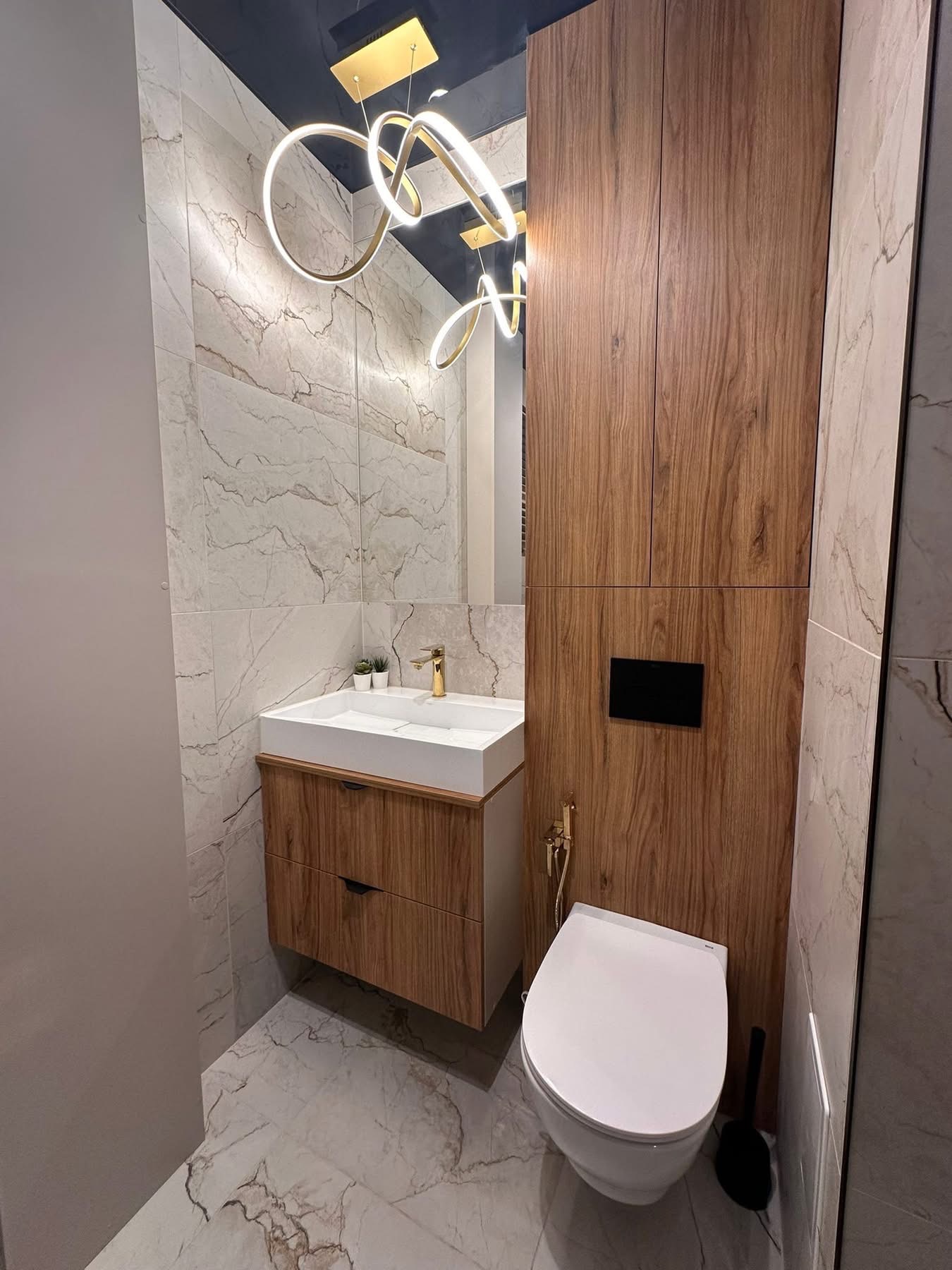 Nowoczesna łazienka z marmurowymi płytkami, drewnianą szafką pod umywalką i designerską lampą sufitową. Uwagę przyciąga złota armatura i ciemny sufit.
