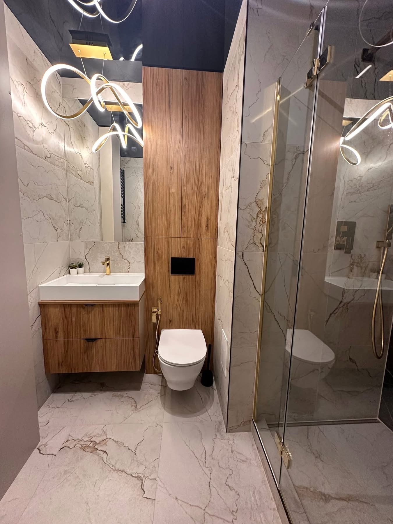 Nowoczesna łazienka z drewnianą zabudową, marmurowymi płytkami i szklaną kabiną prysznicową. Uwagę przykuwa designerska lampa sufitowa.