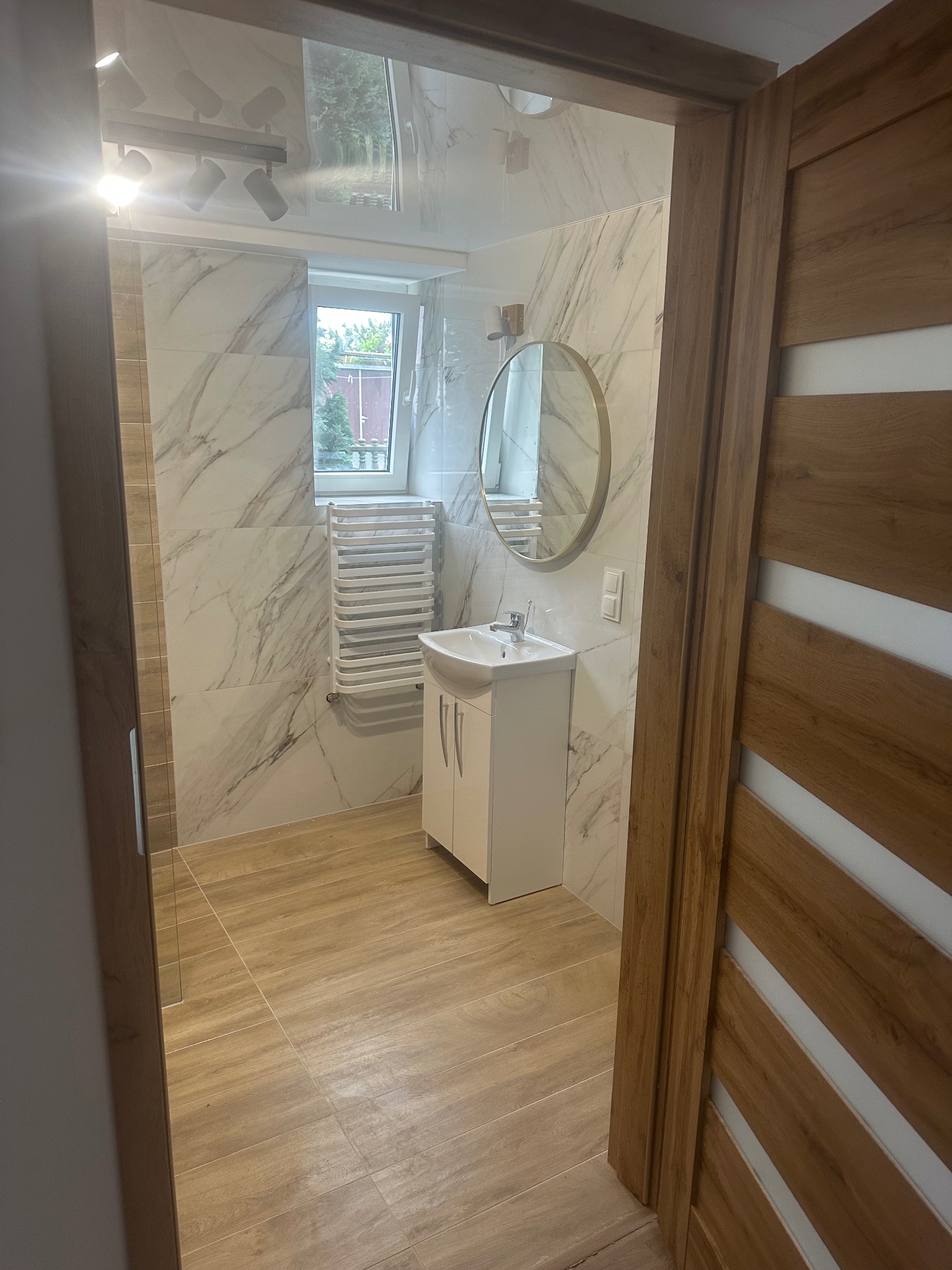 Jasna łazienka z marmurowymi płytkami, białymi meblami i drewnianymi drzwiami. Widoczne oświetlenie sufitowe i okrągłe lustro. Nowoczesny grzejnik drabinkowy.