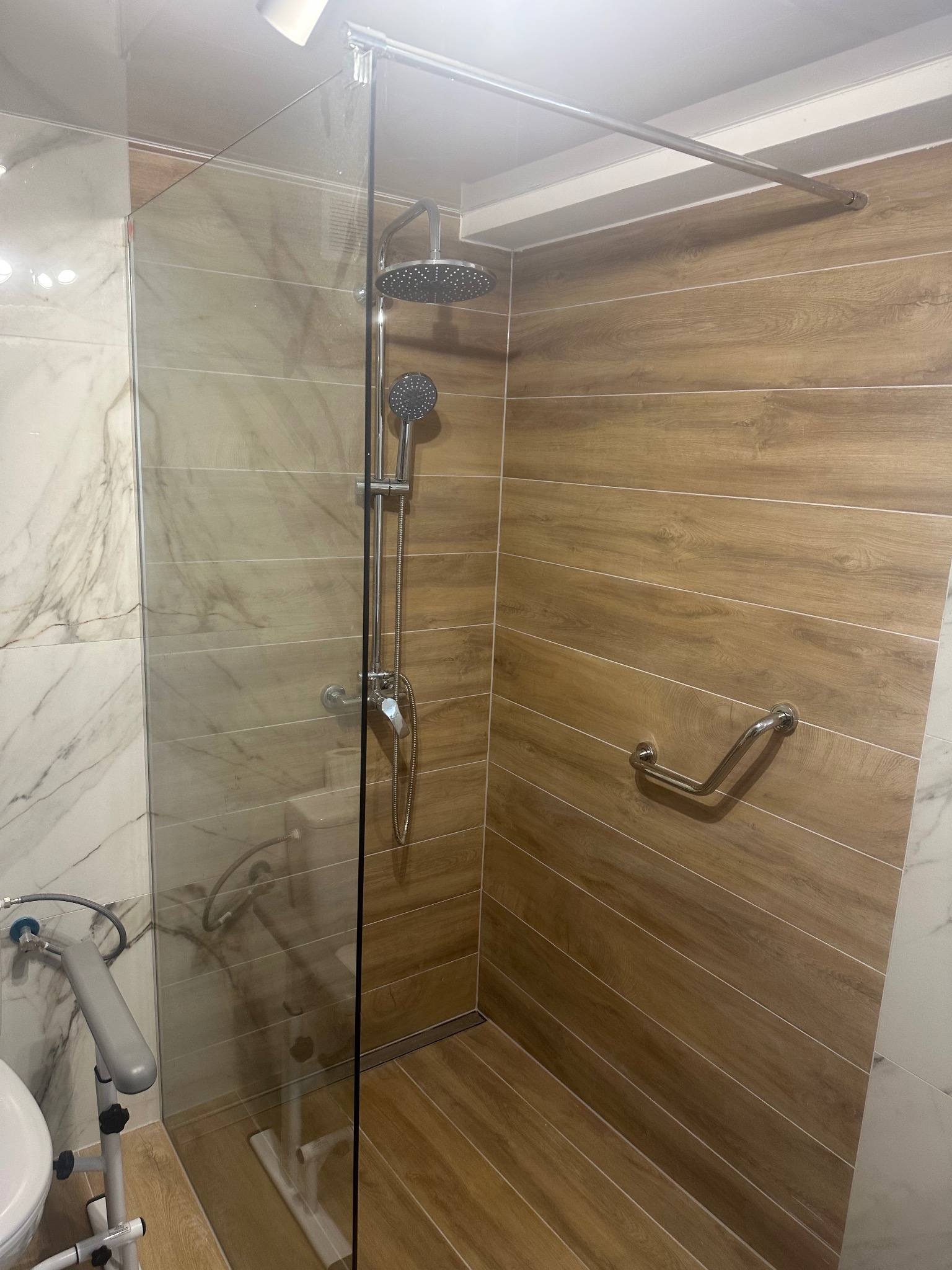 Nowoczesna kabina prysznicowa z deszczownicą i uchwytem dla niepełnosprawnych, wykończona płytkami imitującymi drewno i marmur. Widoczny fragment toalety z podparciem.