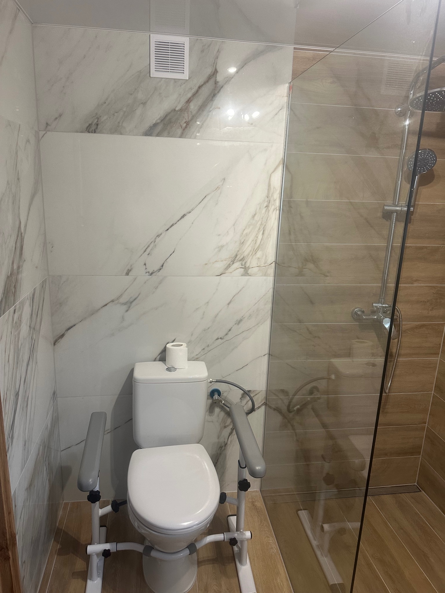Wykończona łazienka z toaletą dla niepełnosprawnych, białe i marmurowe płytki, kabina prysznicowa z drewnopodobnymi płytkami. Widok z góry, estetyczne wykończenie.