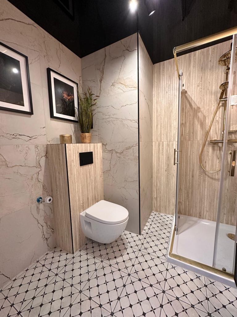 Stylowa łazienka z marmurowymi płytkami, wiszącą toaletą i kabiną prysznicową z złotymi detalami. Czarno-biała podłoga w geometryczny wzór. Nowoczesny design.