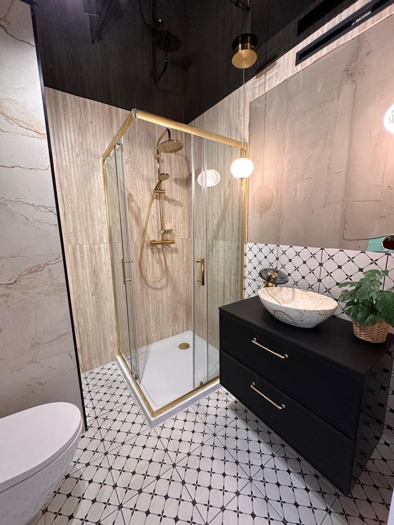 Nowoczesna łazienka z kabiną prysznicową w złotej ramie, czarną szafką z umywalką i wzorzystą podłogą. Minimalistyczny design, oświetlenie punktowe.