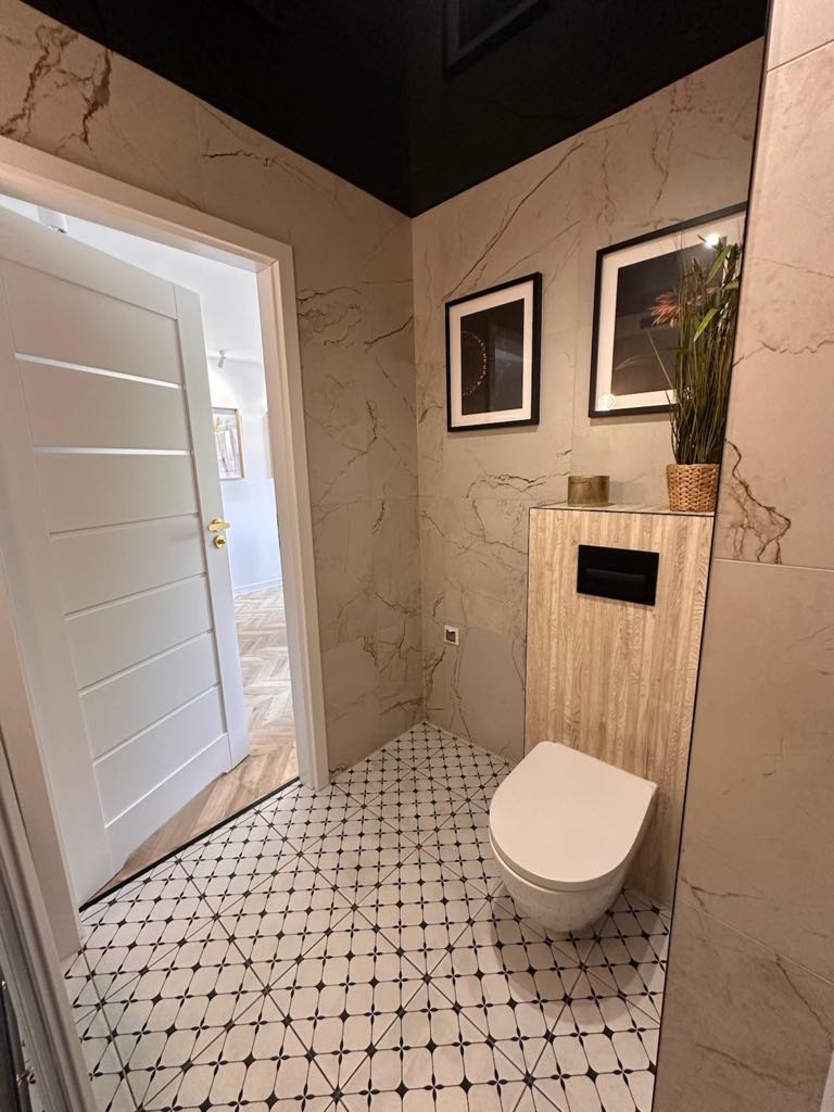 Elegancka łazienka po remoncie: marmurowe płytki, czarno-biała mozaika na podłodze, podwieszana toaleta, czarny sufit i dekoracje ścienne. Widok na otwarte drzwi.
