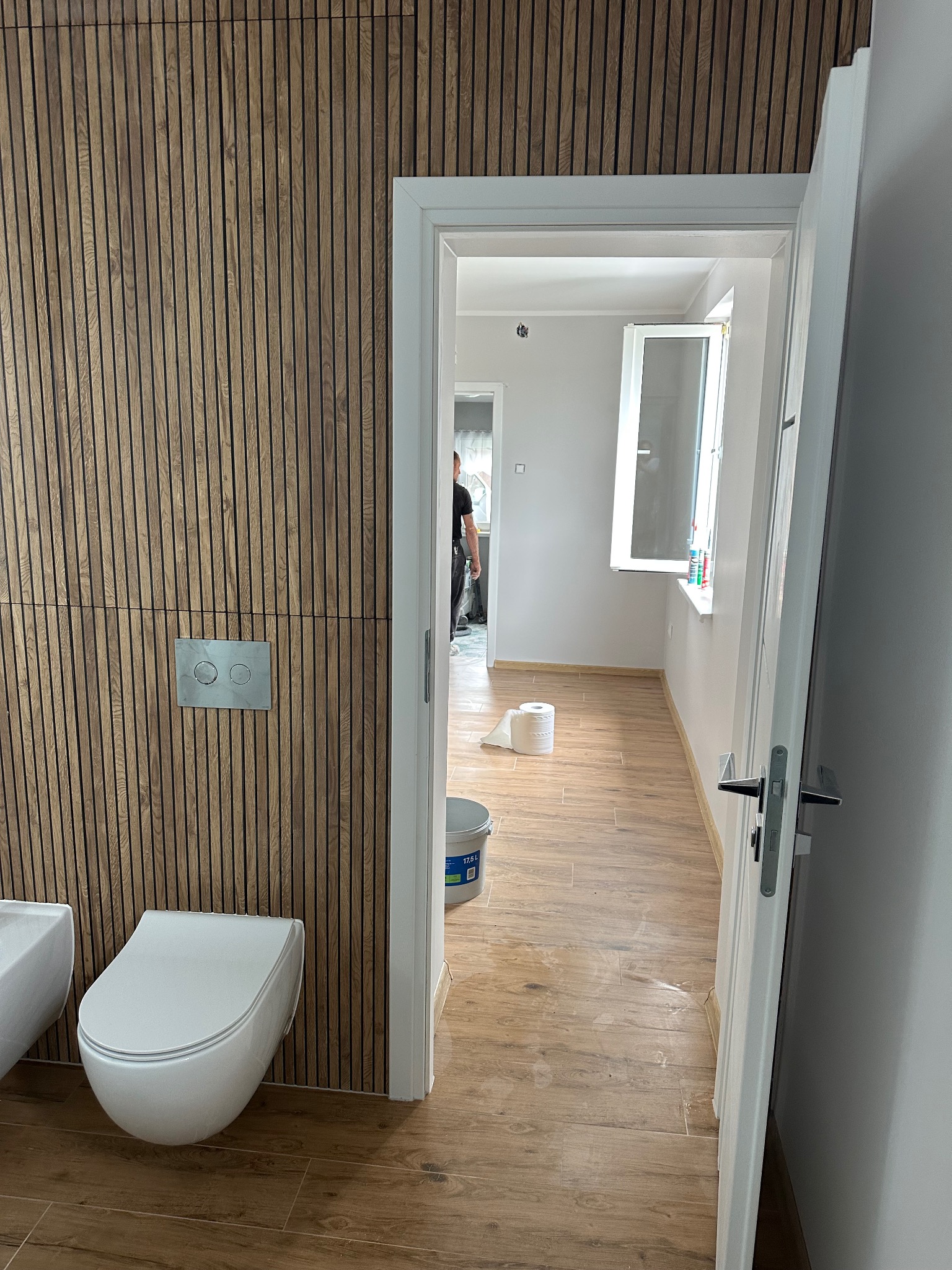 Nowoczesna łazienka po remoncie: drewniane panele ścienne, podwieszana toaleta, płytki imitujące drewno na podłodze. Widok przez otwarte drzwi na dalszą część łazienki.