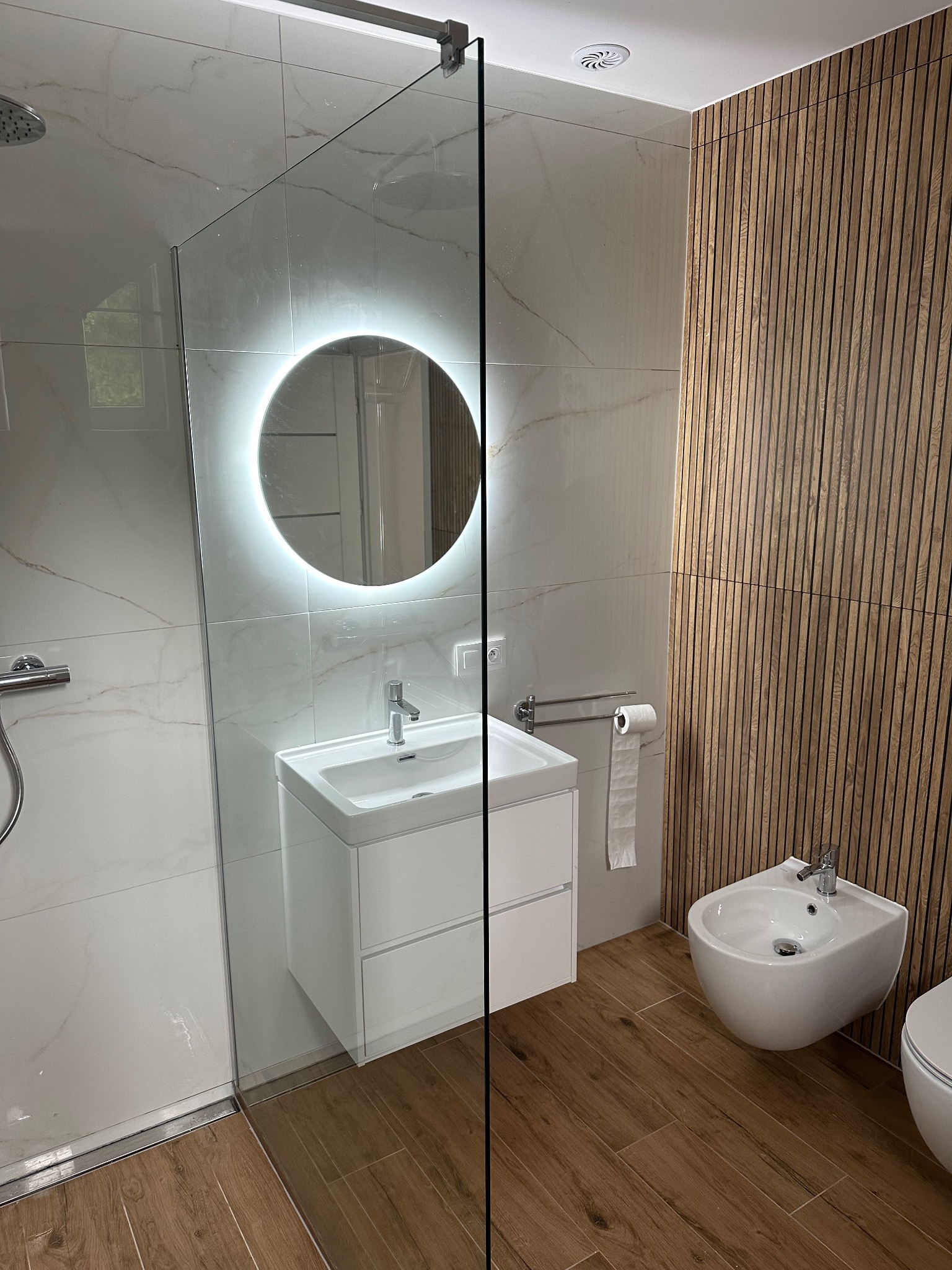 Nowoczesna łazienka po remoncie: biała umywalka z szafką, podświetlane lustro, bidet i deska sedesowa. Ściany wyłożone płytkami imitującymi marmur i drewniane lamele.