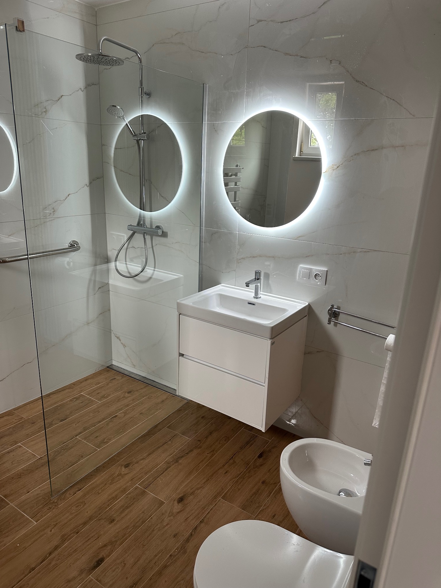 Nowoczesna łazienka z kabiną prysznicową, umywalką z szafką i bidetem. Lustra podświetlane LED. Płytki imitujące marmur i drewnopodobna podłoga.