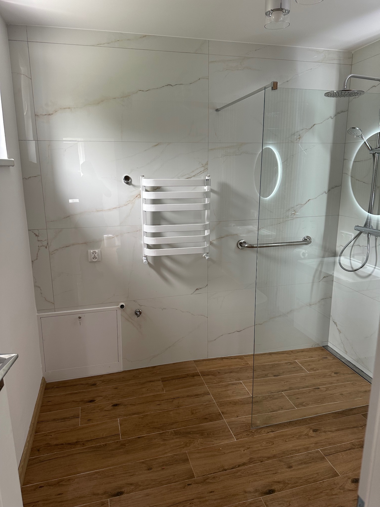 Nowoczesna łazienka z kabiną walk-in, marmurowe płytki ścienne, drewnopodobne płytki podłogowe, grzejnik drabinkowy i okrągłe lustro z podświetleniem LED.