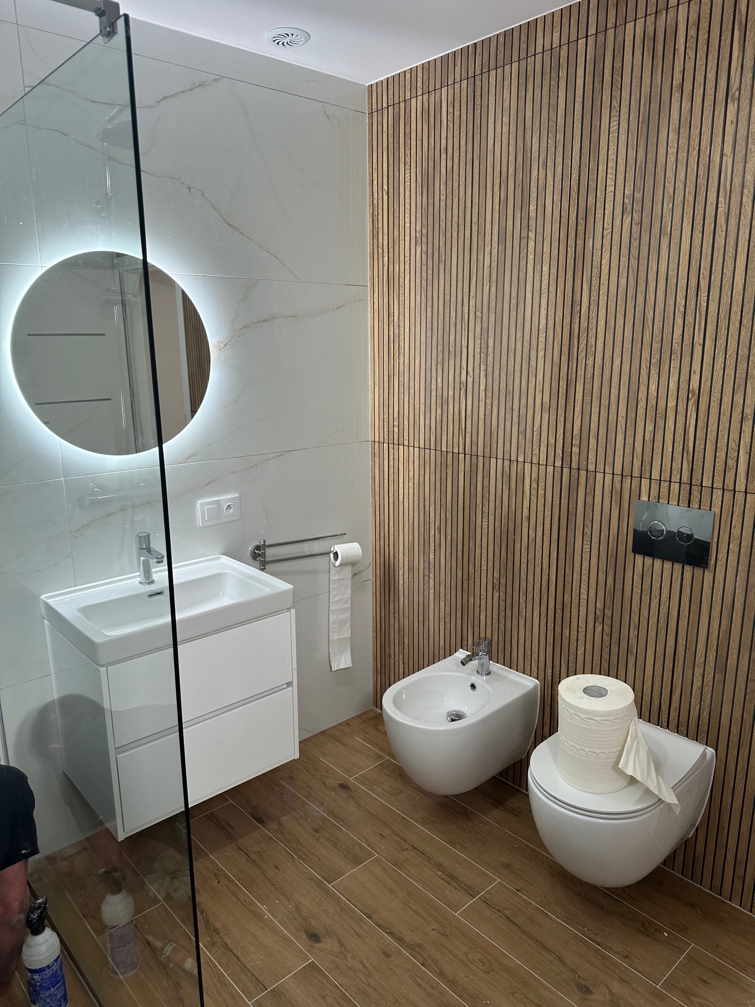 Nowoczesna łazienka z białymi płytkami imitującymi marmur, drewnianą ścianą z lameli, okrągłym lustrem z podświetleniem LED i podłogą z płytek imitujących drewno.