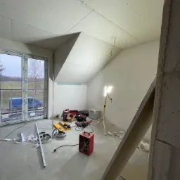 Poddasze w trakcie wykańczania, widoczne płyty na skośnym suficie, narzędzia rozłożone na podłodze, okno z widokiem na zewnątrz i grzejnik elektryczny. Oświetlenie robocze skierowane na ścianę.