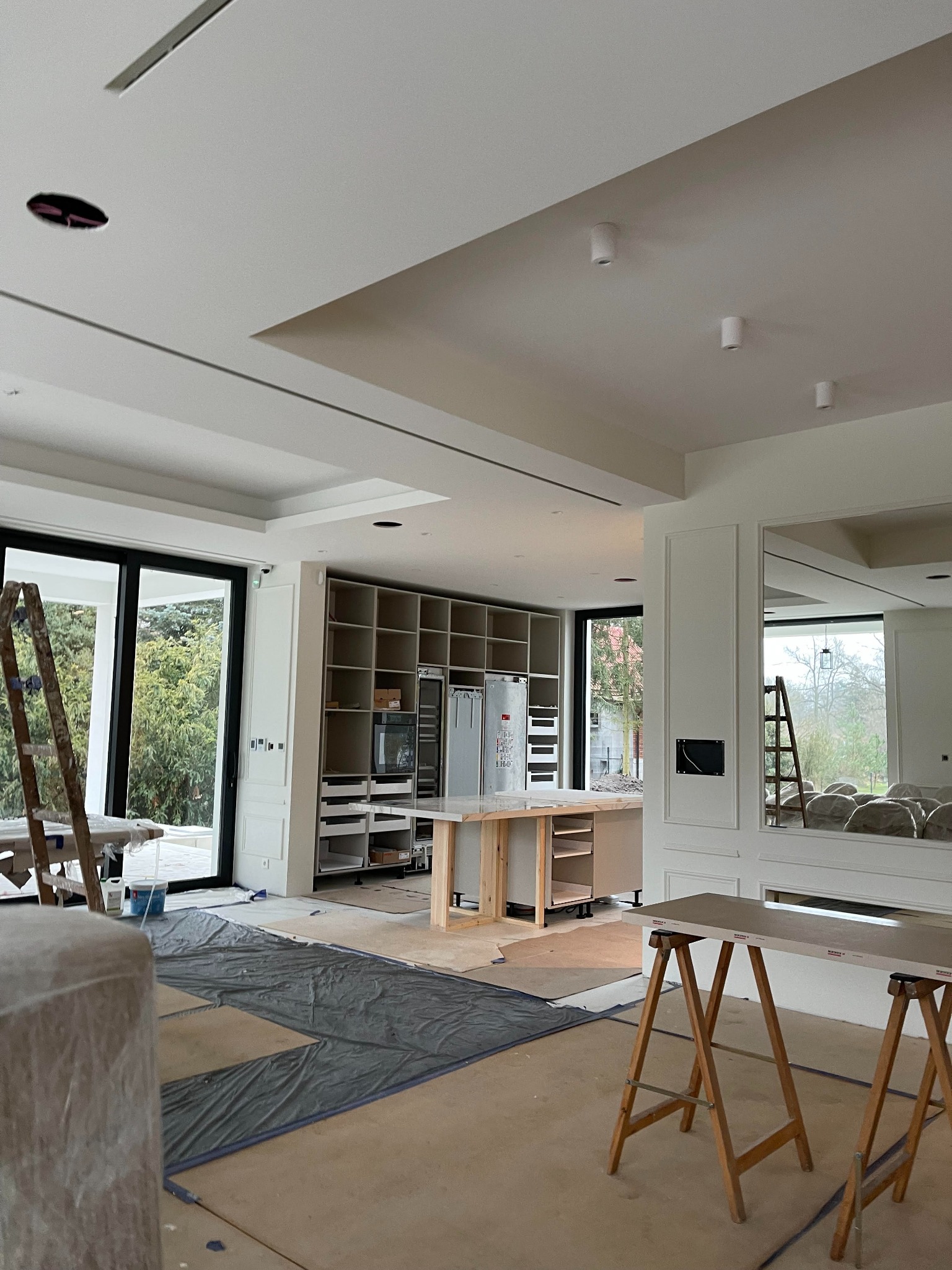 Wnętrze domu w trakcie wykończenia, widoczne zabudowy meblowe kuchenne, przygotowane do montażu, pomalowane ściany i sufit, zabezpieczona podłoga, drabiny i kozły malarskie.