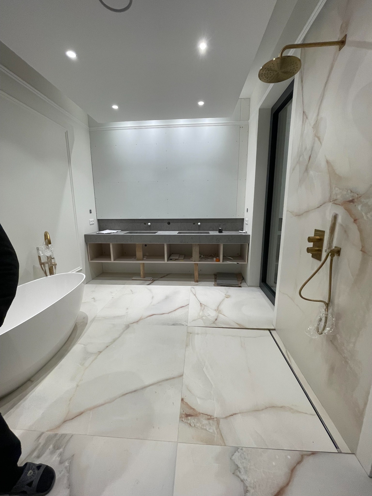 Wnętrze łazienki w trakcie remontu: biała wanna wolnostojąca, marmurowe płytki podłogowe i ścienne, surowy betonowy blat pod umywalki z otwartymi szafkami pod spodem, złota deszczownica i bateria...