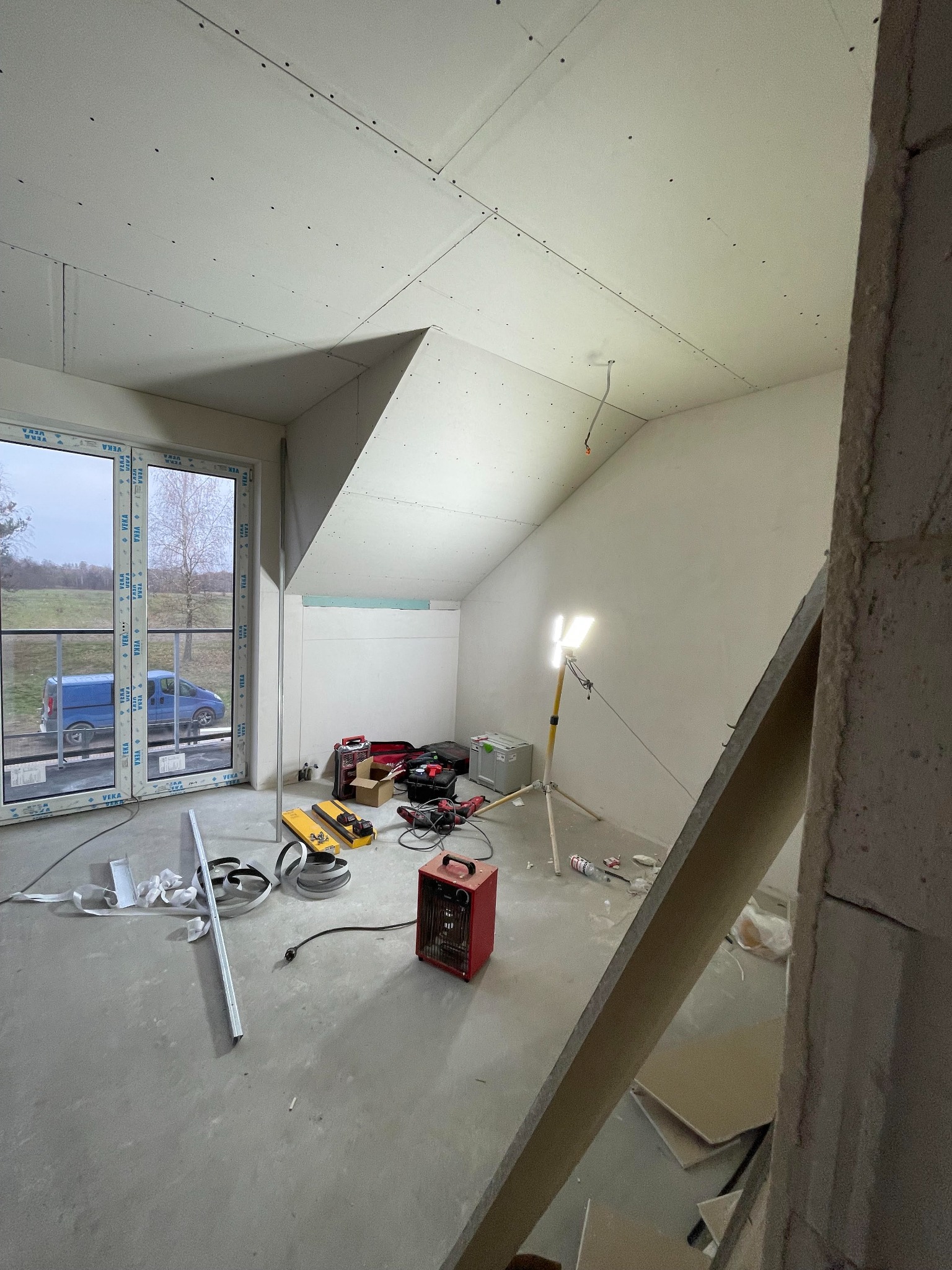 Poddasze w trakcie wykańczania, widoczne płyty na skośnym suficie, narzędzia rozłożone na podłodze, okno z widokiem na zewnątrz i grzejnik elektryczny. Oświetlenie robocze skierowane na ścianę.