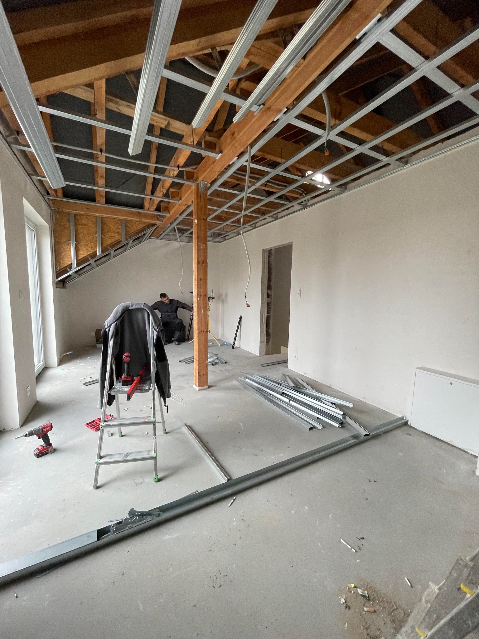 Wnętrze w trakcie montażu stelaża sufitu podwieszanego z profili metalowych, widoczna konstrukcja drewniana dachu, drabina z narzędziami i pracownik w tle.