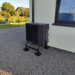 Czarna jednostka zewnętrzna pompy ciepła Panasonic Aquarea zamontowana na metalowej podstawie z widocznymi przewodami na elewacji domu z szarą elewacją i oknem w czarnej ramie.