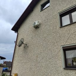 KLIMADAM Adam Skorupa - Boczna ściana domu z widoczną jednostką zewnętrzną klimatyzatora, anteną satelitarną i oknami z brązowymi ramami, na tle pochmurnego nieba.