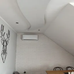 Biały klimatyzator ścienny zamontowany nad ceglaną ścianą w jasnym pokoju na poddaszu, obok dekoracyjna metalowa głowa jelenia.