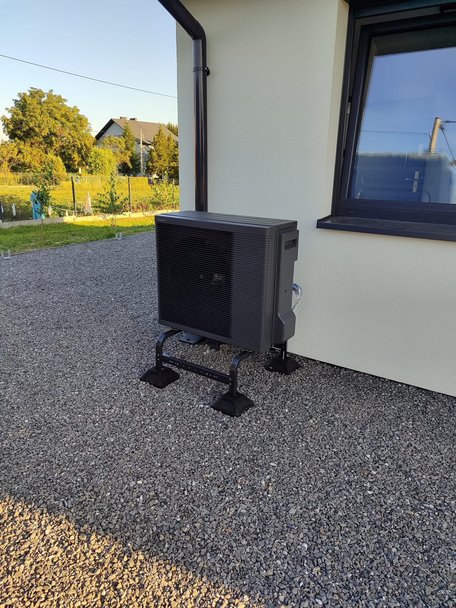 Czarna jednostka zewnętrzna pompy ciepła Panasonic Aquarea zamontowana na metalowej podstawie z widocznymi przewodami na elewacji domu z szarą elewacją i oknem w czarnej ramie.