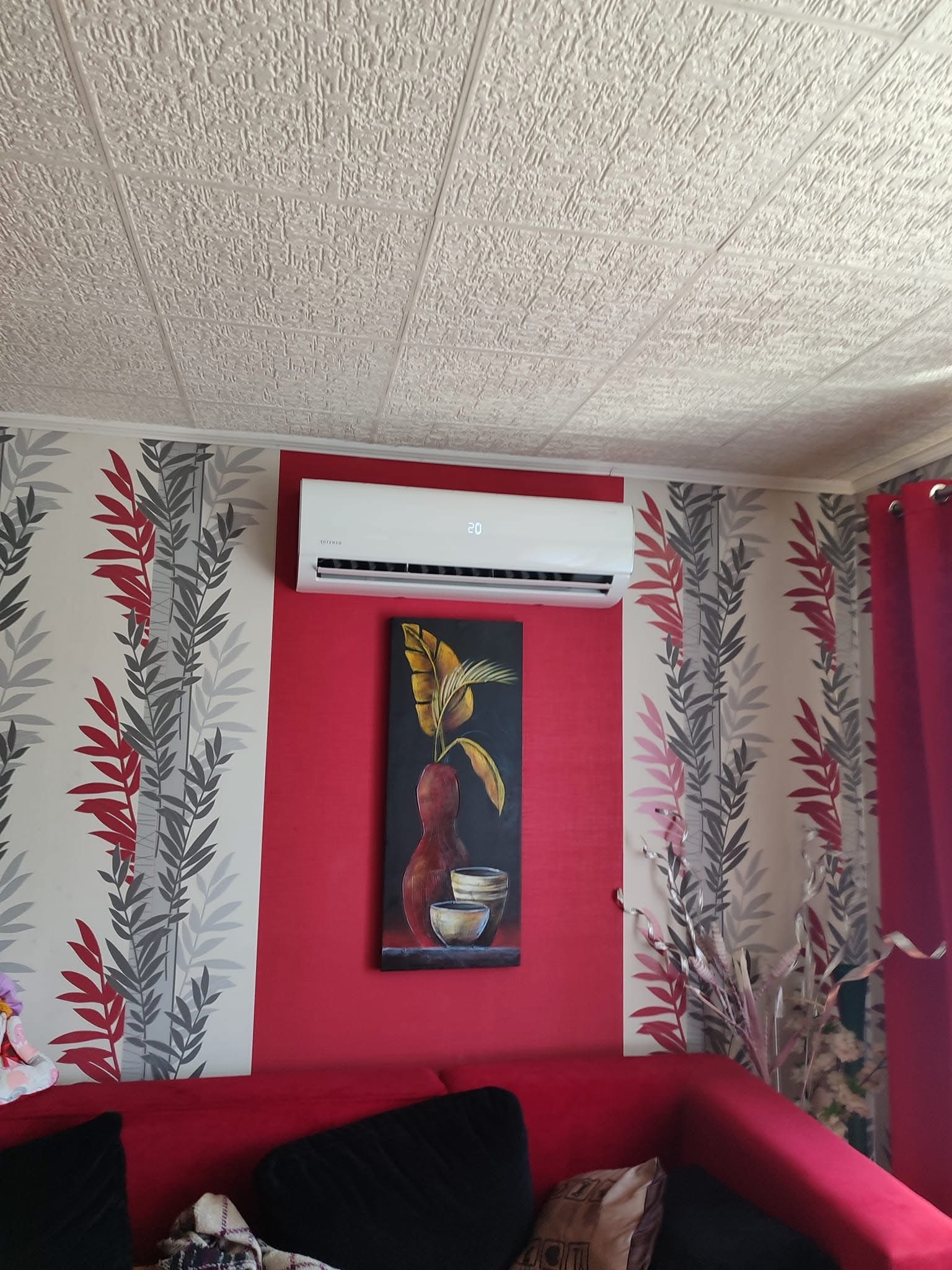 Biała jednostka klimatyzacyjna zamontowana na czerwonej ścianie nad obrazem z motywem roślinnym, widoczna w salonie z wzorzystą tapetą i czerwonymi zasłonami.