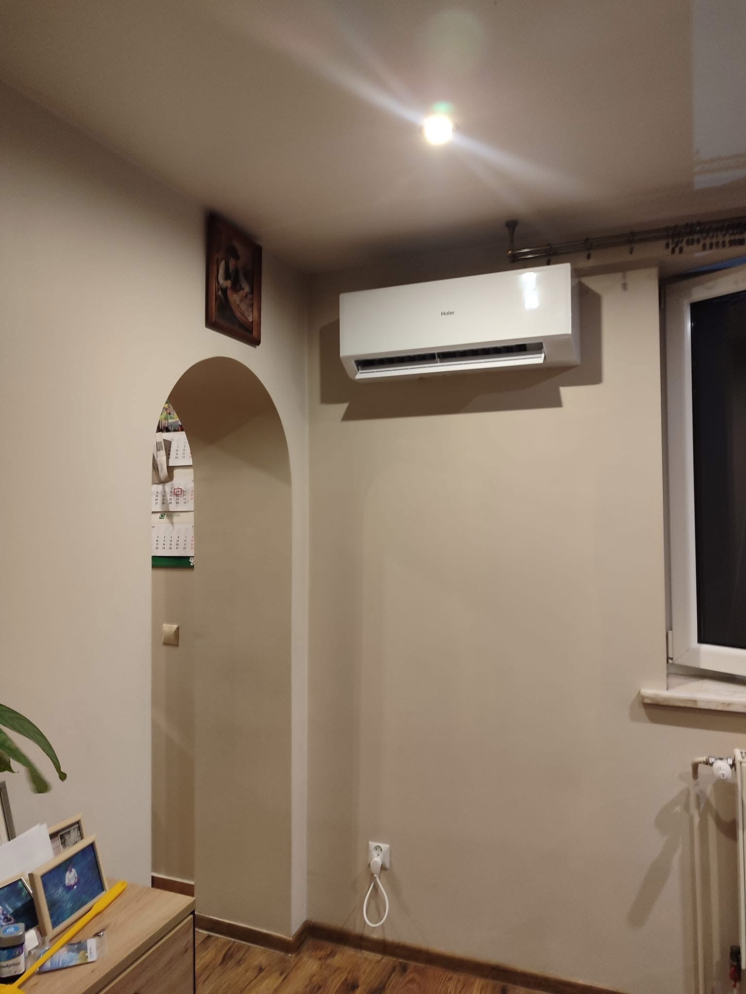 Biała klimatyzacja Haier zamontowana na ścianie w pokoju z drewnianą podłogą, widoczny kalendarz w przejściu łukowym i fragment okna.