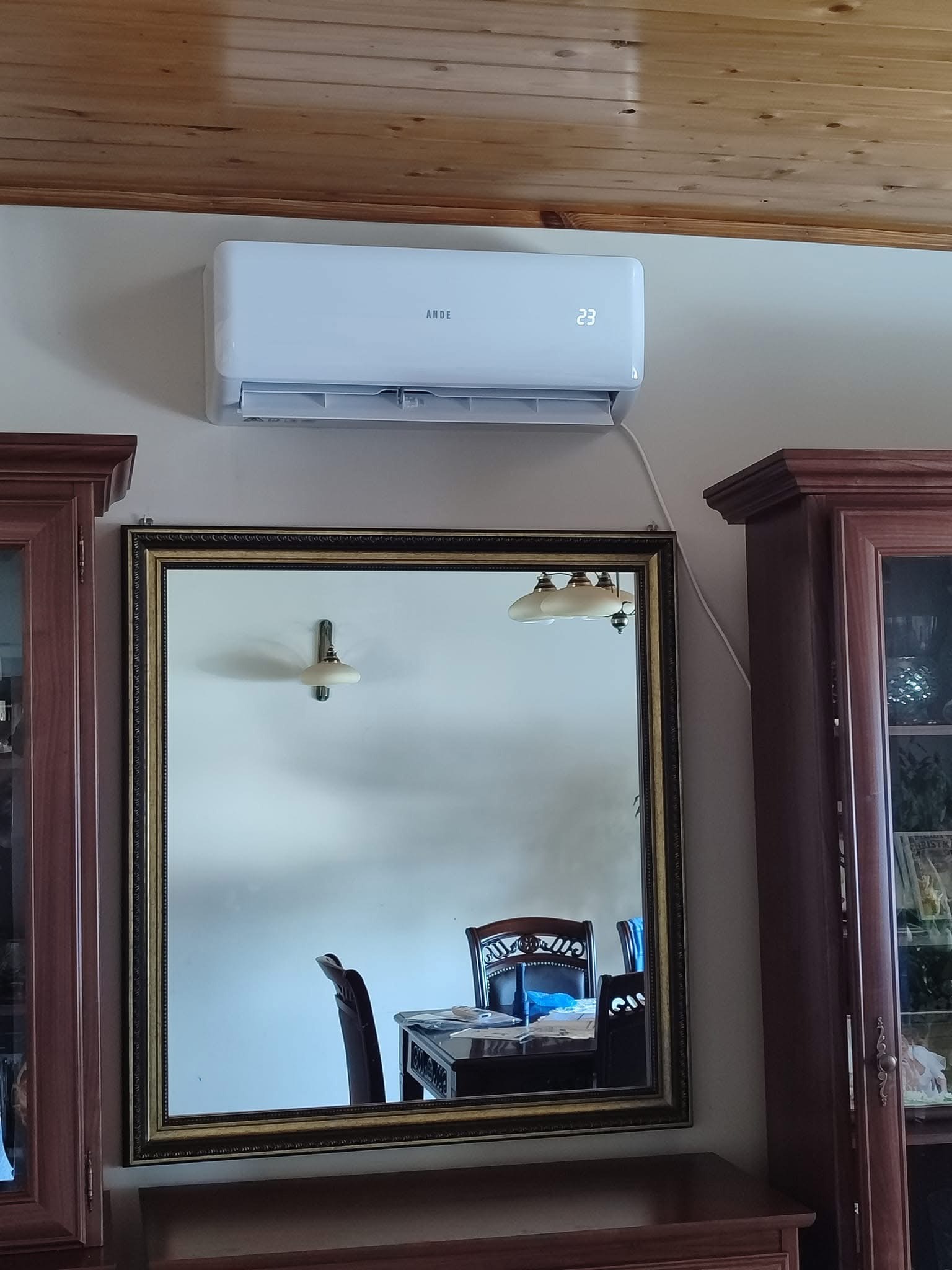 Biały klimatyzator marki ANDE zamontowany na ścianie nad lustrem w złotej ramie, odbijającym stół i krzesła w jadalni z drewnianym sufitem.