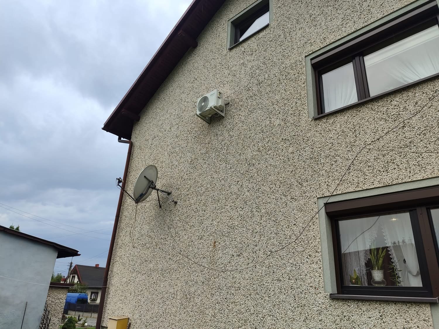 Boczna ściana domu z widoczną jednostką zewnętrzną klimatyzatora, anteną satelitarną i oknami z brązowymi ramami, na tle pochmurnego nieba.