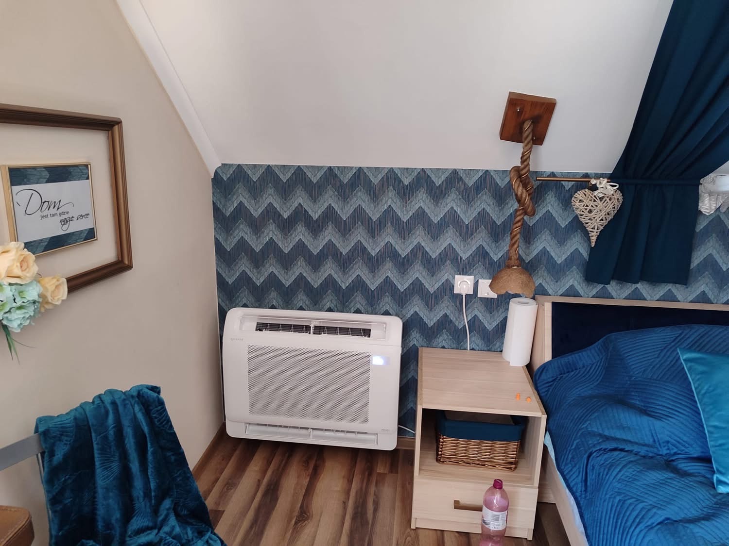 Klimatyzator podłogowy w sypialni z niebieskimi akcentami, widoczny na tle ściany w zygzaki, obok szafki nocnej z dekoracyjną lampą i zasłonami w podobnej kolorystyce.