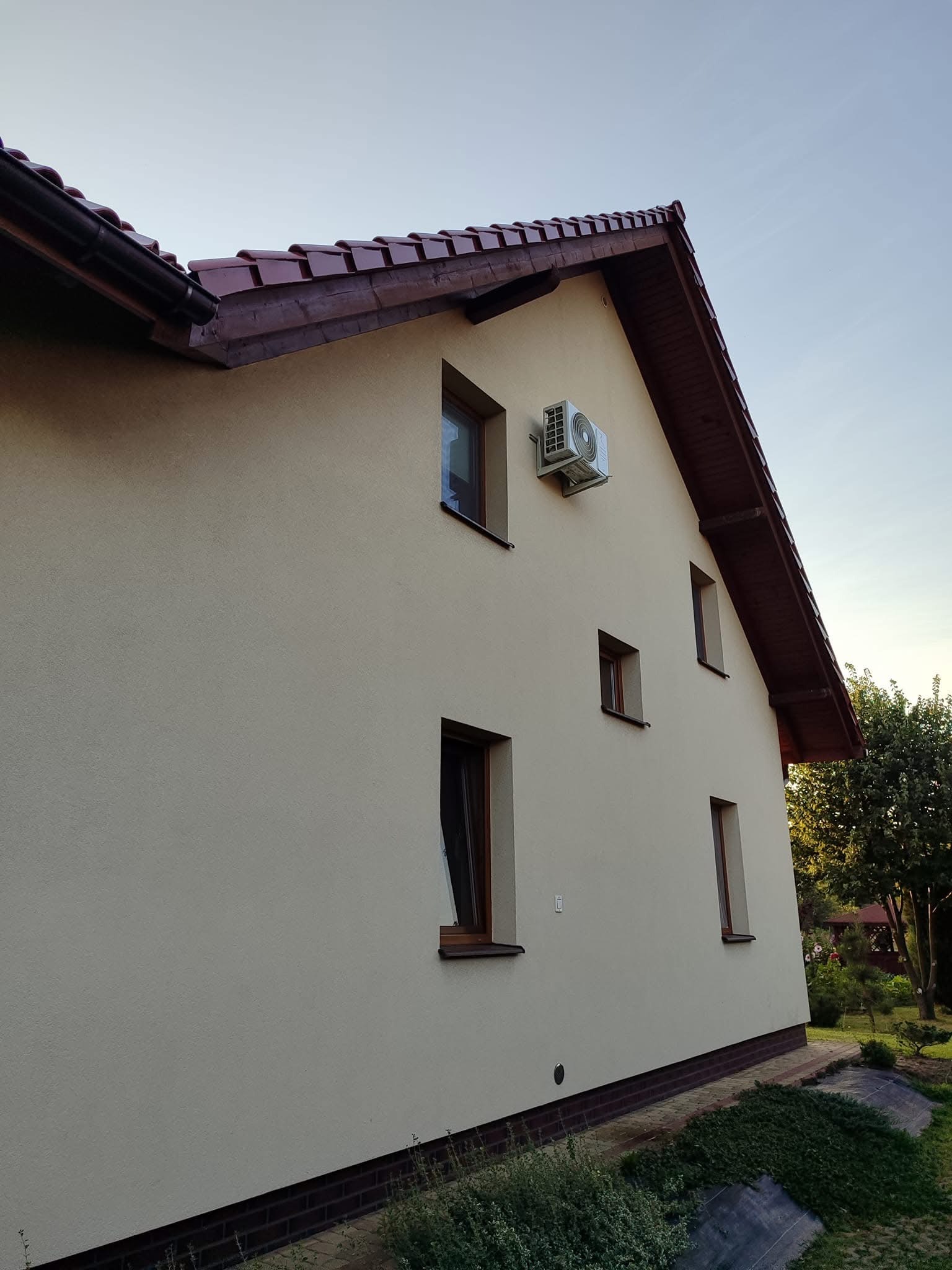 Zewnętrzna jednostka klimatyzatora zamontowana na elewacji domu jednorodzinnego z brązową dachówką i drewnianymi oknami. Widok z boku, w tle zieleń ogrodu.