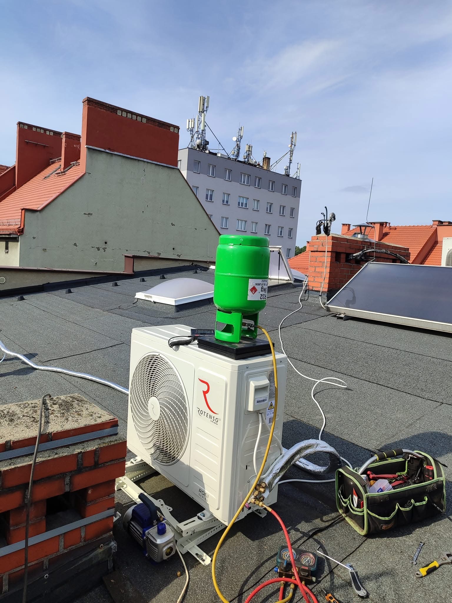 Montaż klimatyzacji Rotenso na dachu budynku, widoczna butla z czynnikiem chłodniczym R32, pompa próżniowa, manometry, narzędzia i skrzynka narzędziowa.