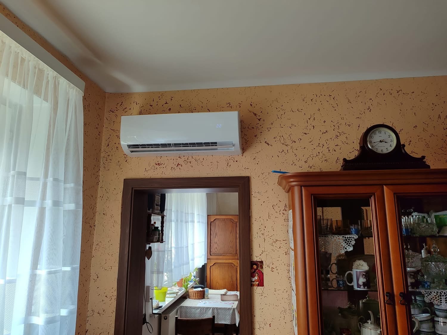 Biały klimatyzator ścienny zamontowany w salonie z beżowymi ścianami o nieregularnej fakturze, widoczne firanki w oknie i stary zegar na kredensie.