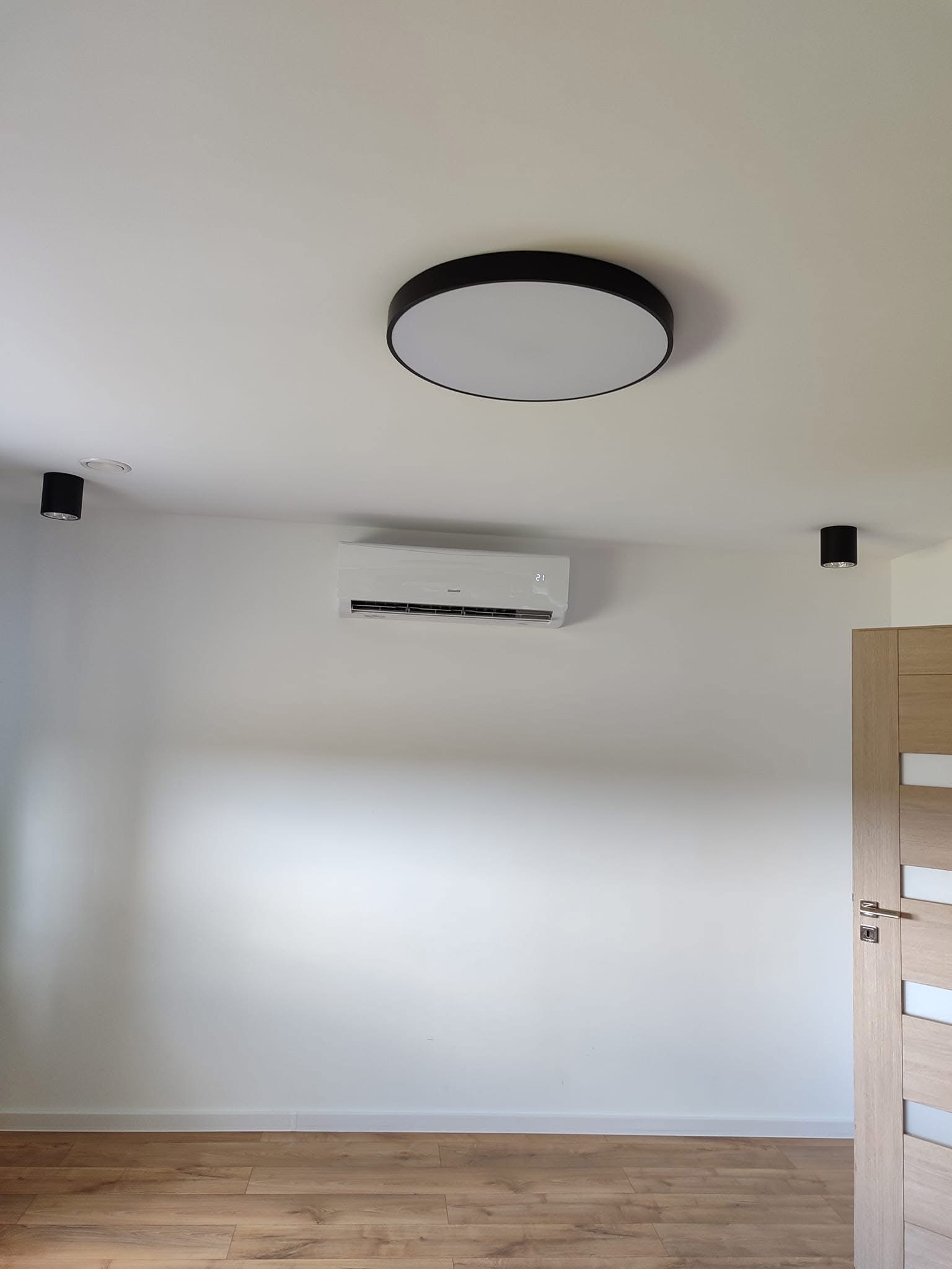 Biały klimatyzator na ścianie w jasnym pomieszczeniu z drewnianą podłogą, widoczne okrągłe lampy sufitowe i fragment drzwi.