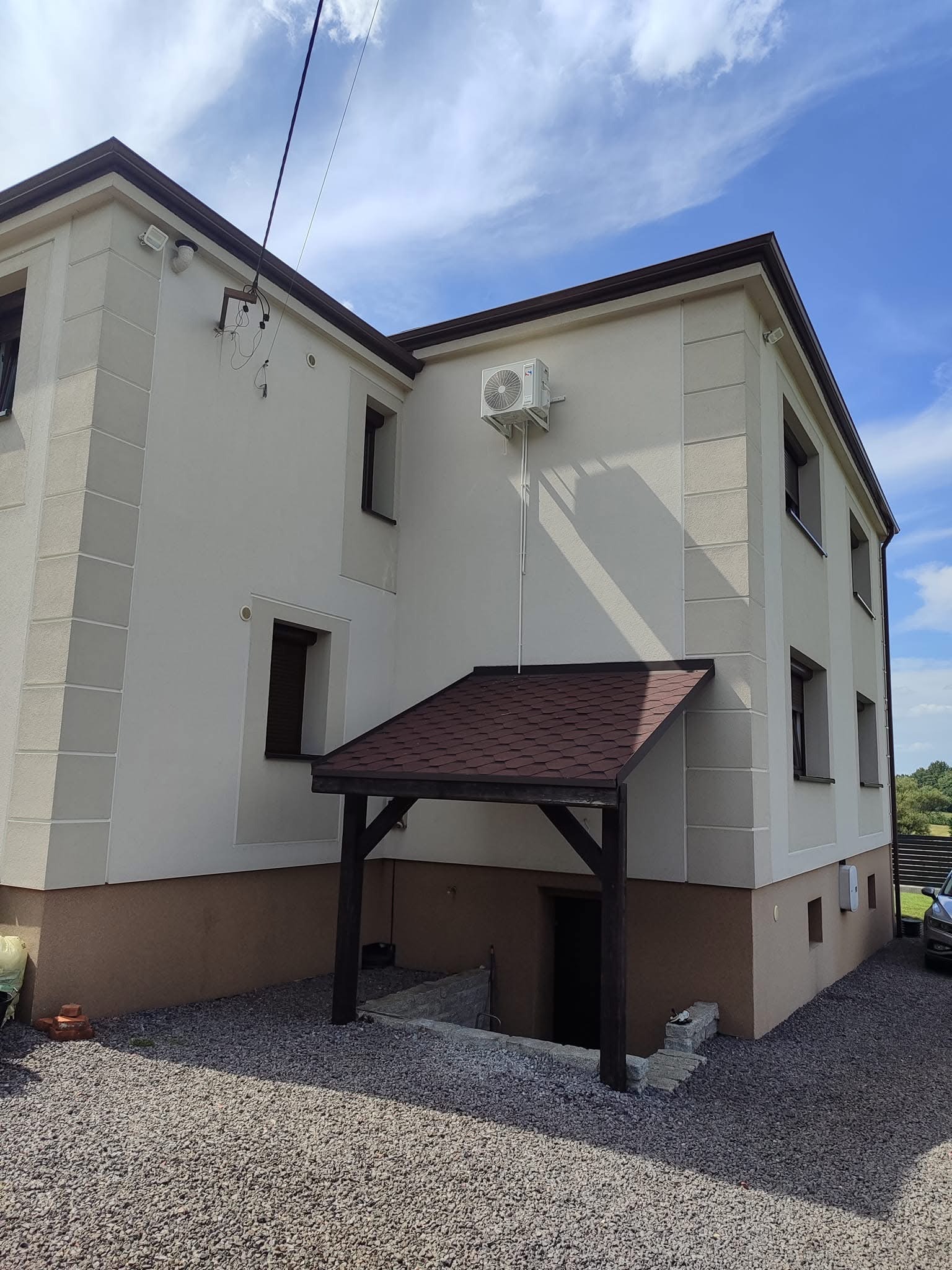 Zewnętrzna jednostka klimatyzacji zamontowana na elewacji dwupiętrowego domu z brązowym zadaszeniem nad wejściem do piwnicy, widok z boku na tle błękitnego nieba.