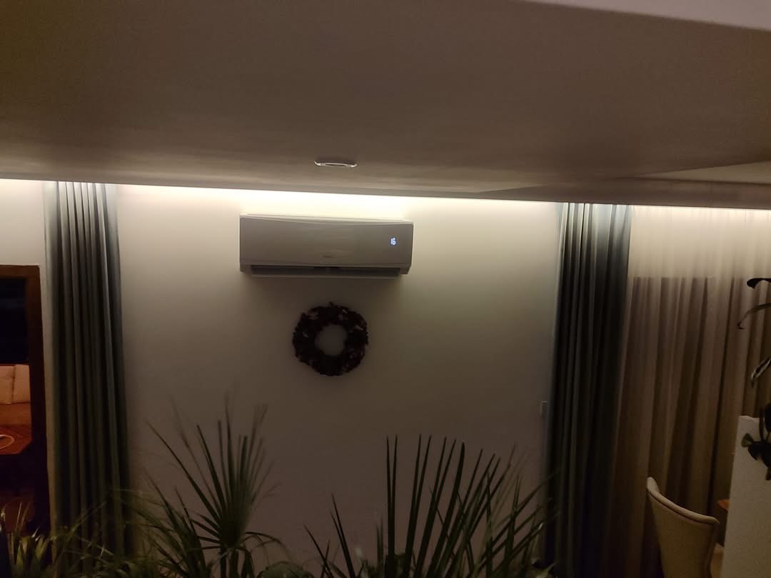 Nowoczesny klimatyzator ścienny w salonie, widoczny wyświetlacz temperatury, dekoracyjny wieniec na ścianie, oświetlenie LED podsufitowe, zasłony w odcieniach szarości, rośliny doniczkowe w dolnej...