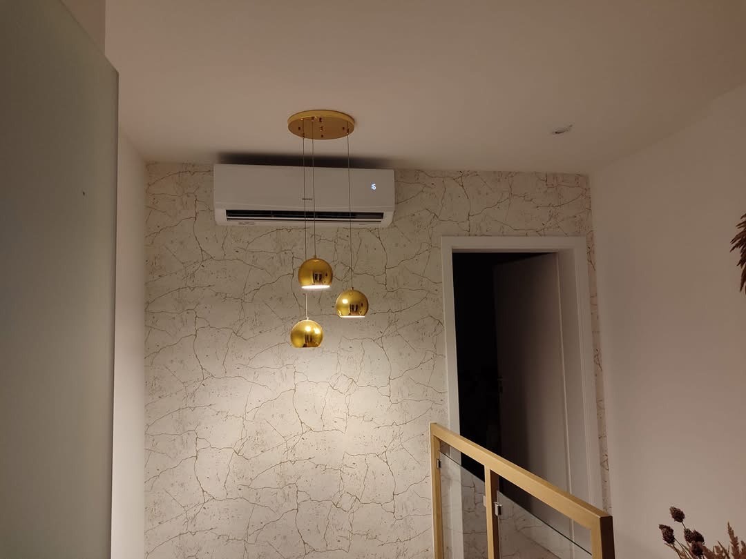 Biała jednostka klimatyzacyjna zamontowana na ścianie z wzorem imitującym spękania, oświetlona przez złotą lampę wiszącą z trzema kulistymi kloszami.