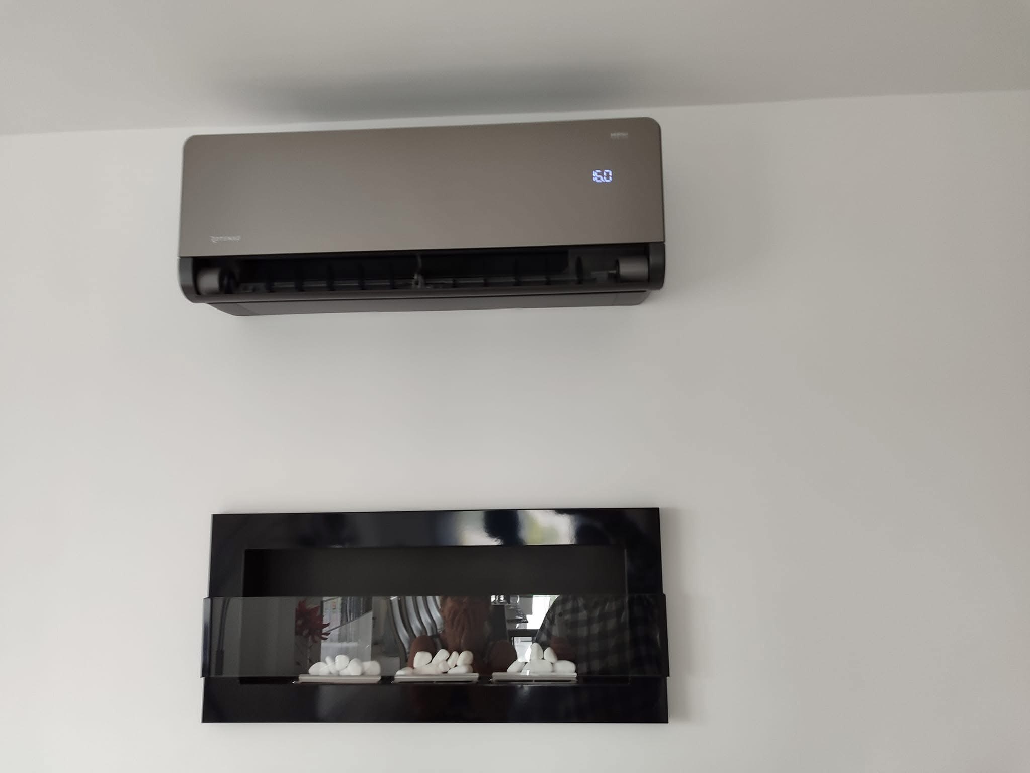 Szara klimatyzacja ścienna z wyświetlaczem temperatury (16 stopni Celsjusza) zamontowana nad czarnym, dekoracyjnym kominkiem elektrycznym z białymi kamykami.