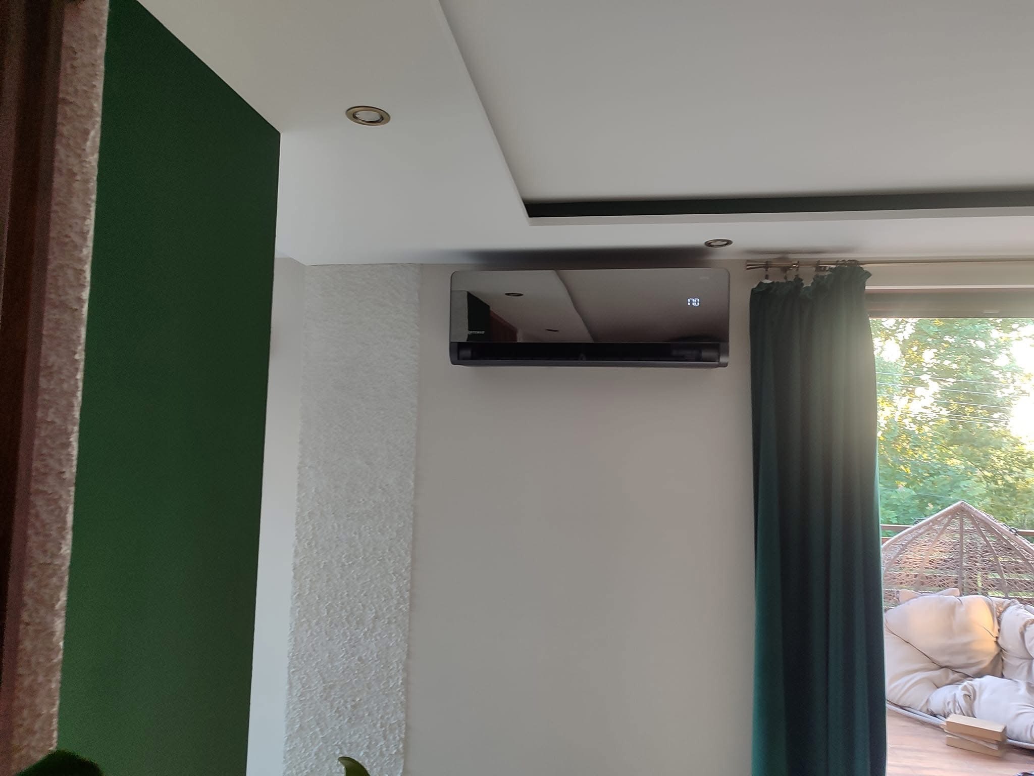 Czarny klimatyzator ścienny z wyświetlaczem temperatury (23 stopnie Celsjusza) w nowoczesnym salonie z zieloną ścianą i widokiem na zewnątrz przez okno zasłonięte ciemnozieloną zasłoną.