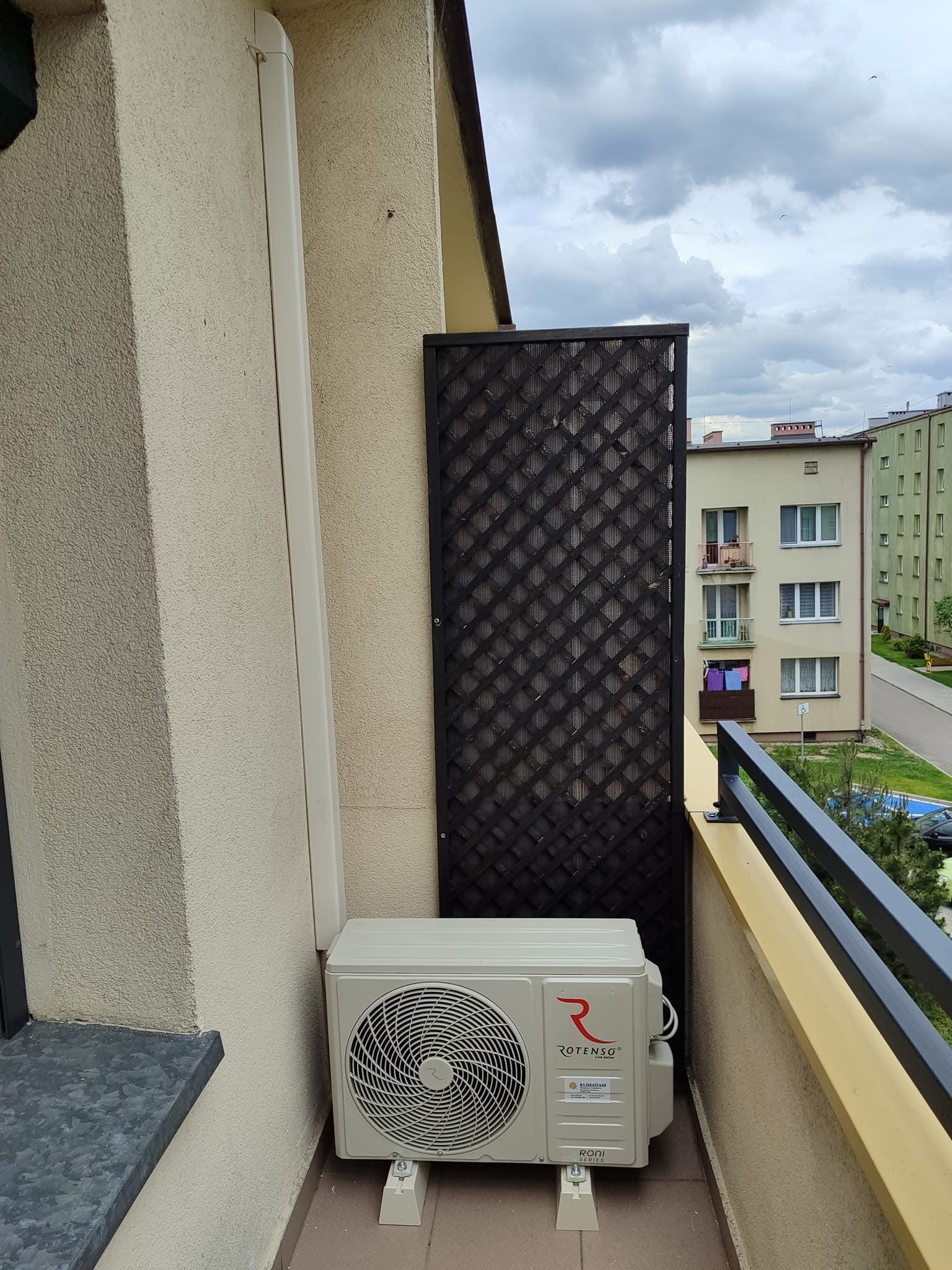 Zewnętrzna jednostka klimatyzatora marki Rotenso zamontowana na balkonie bloku mieszkalnego, widok na siatkę balkonową i zabudowę miejską w tle.
