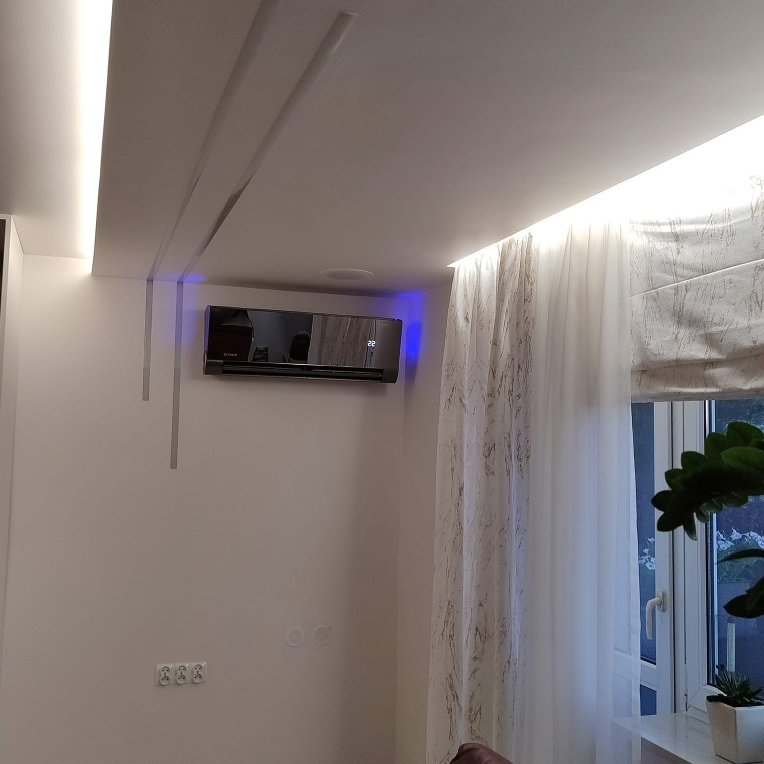 Czarny klimatyzator ścienny z niebieskim podświetleniem, zamontowany w nowoczesnym wnętrzu z dekoracyjnym oświetleniem sufitowym i zasłonami w marmurkowy wzór.