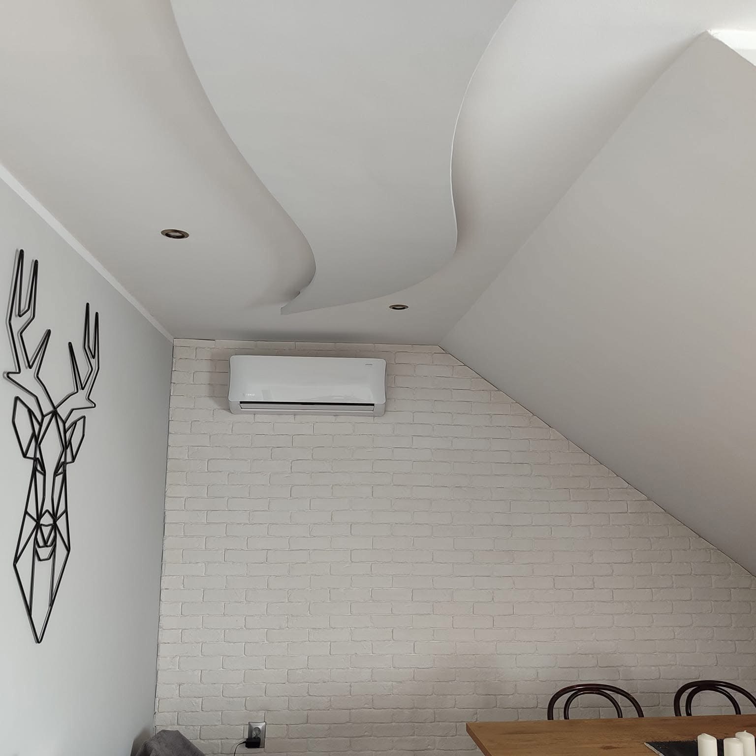Biały klimatyzator ścienny zamontowany nad ceglaną ścianą w jasnym pokoju na poddaszu, obok dekoracyjna metalowa głowa jelenia.