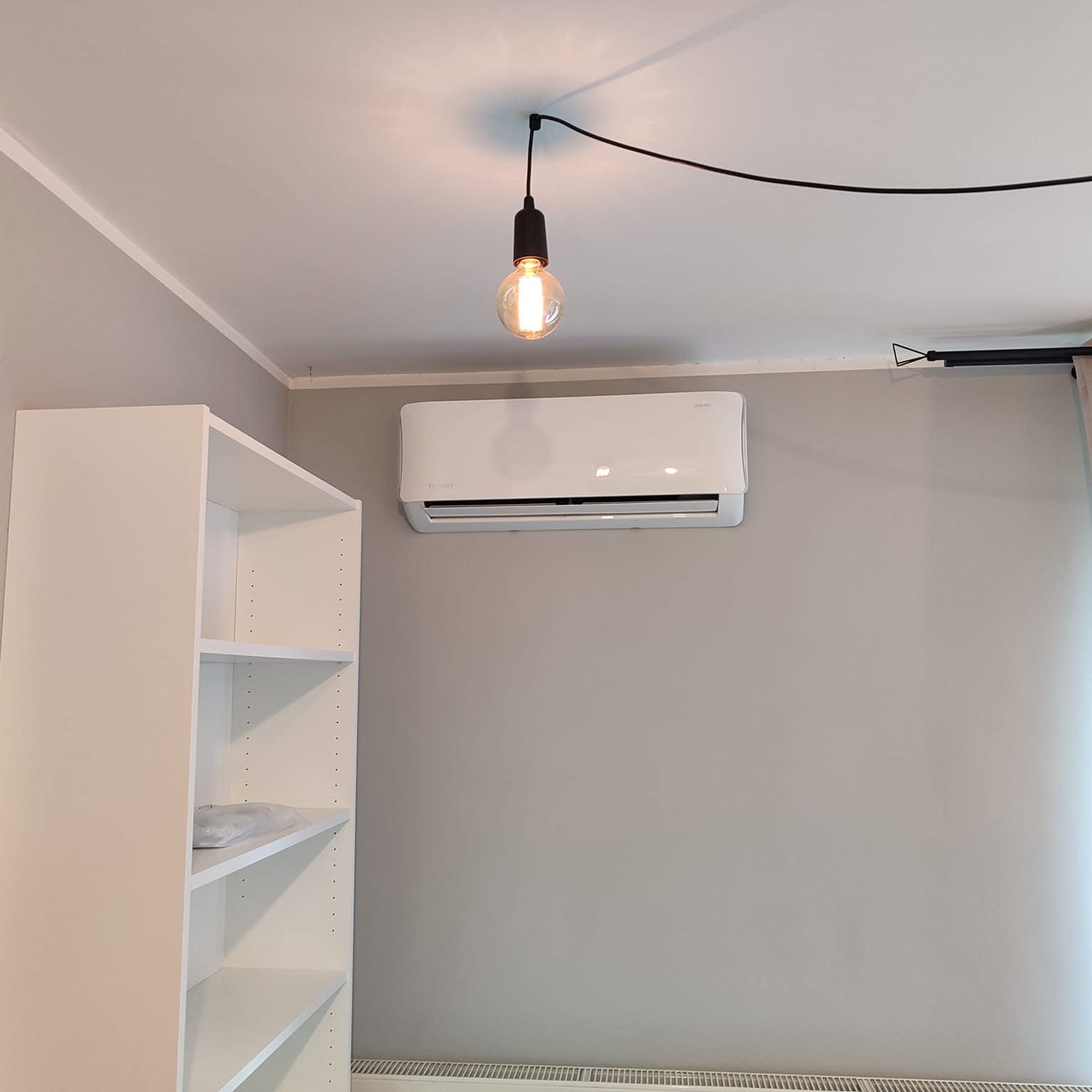 Biały klimatyzator ścienny zamontowany w szarym pokoju, oświetlony żarówką Edisona zwisającą z czarnego kabla, obok biały regał z pustymi półkami.