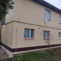 Dwukondygnacyjny budynek po nałożeniu jasnobeżowej warstwy elewacji, okna zabezpieczone folią, widoczny cokół w kolorze ciemnego brązu, na ścianie numer domu '6'.