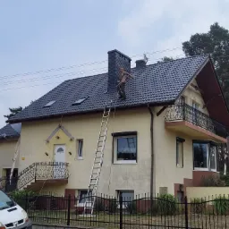 Pracownik w uprzęży na dachu domu z ciemną dachówką i kominem z cegły, po malowaniu, drabina oparta o dach, widok z dołu, elewacja domu w jasnym kolorze.