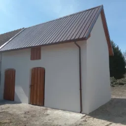 Świeżo otynkowany budynek gospodarczy z brązowym, metalowym dachem i drewnianymi drzwiami w rustykalnym stylu, widoczny fragment starego budynku z anteną satelitarną w tle.