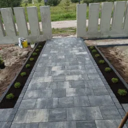 Ścieżka z szarej kostki brukowej prowadząca do betonowego ogrodzenia z charakterystycznymi prześwitami, obsadzona po bokach młodymi roślinami w świeżej ziemi.