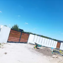 Nowoczesne ogrodzenie z białych betonowych przęseł i drewnianych paneli bramy przesuwnej, widok w trakcie prac ziemnych z taczką na pierwszym planie.