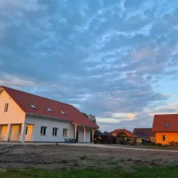 Nowo wybudowany, parterowy dom z białymi ścianami i czerwoną dachówką w otoczeniu innych budynków i terenu przygotowanego pod ogród, uchwycony w świetle zachodzącego słońca.