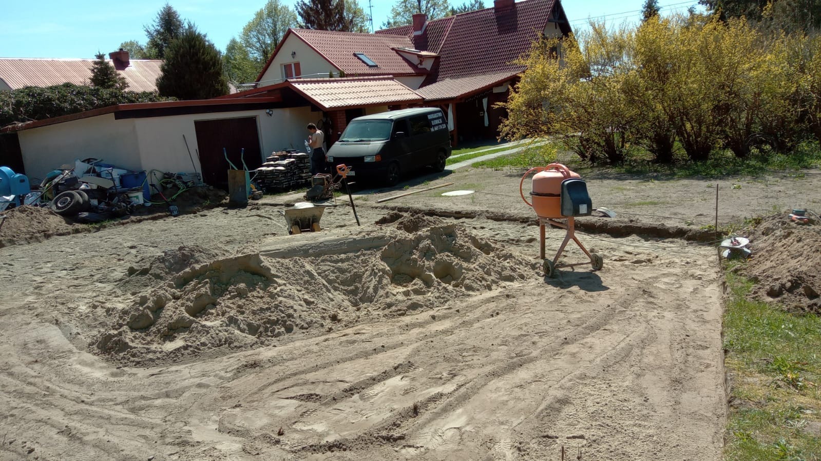 Przygotowanie podłoża pod przyszłą nawierzchnię: wyrównany piasek, widoczna linia wykopu, betoniarka, taczka, zaparkowany bus firmy budowlanej i sterty kostki brukowej w tle.