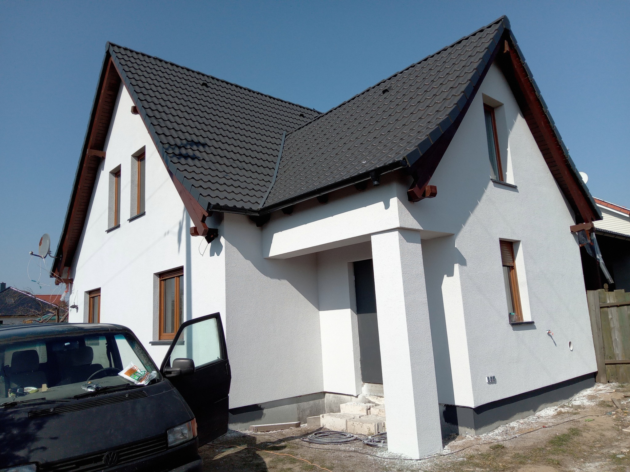 Nowo otynkowany dom jednorodzinny z czarną dachówką i otwartymi drzwiami czarnego samochodu dostawczego, widoczne ślady prac wykończeniowych wokół budynku.