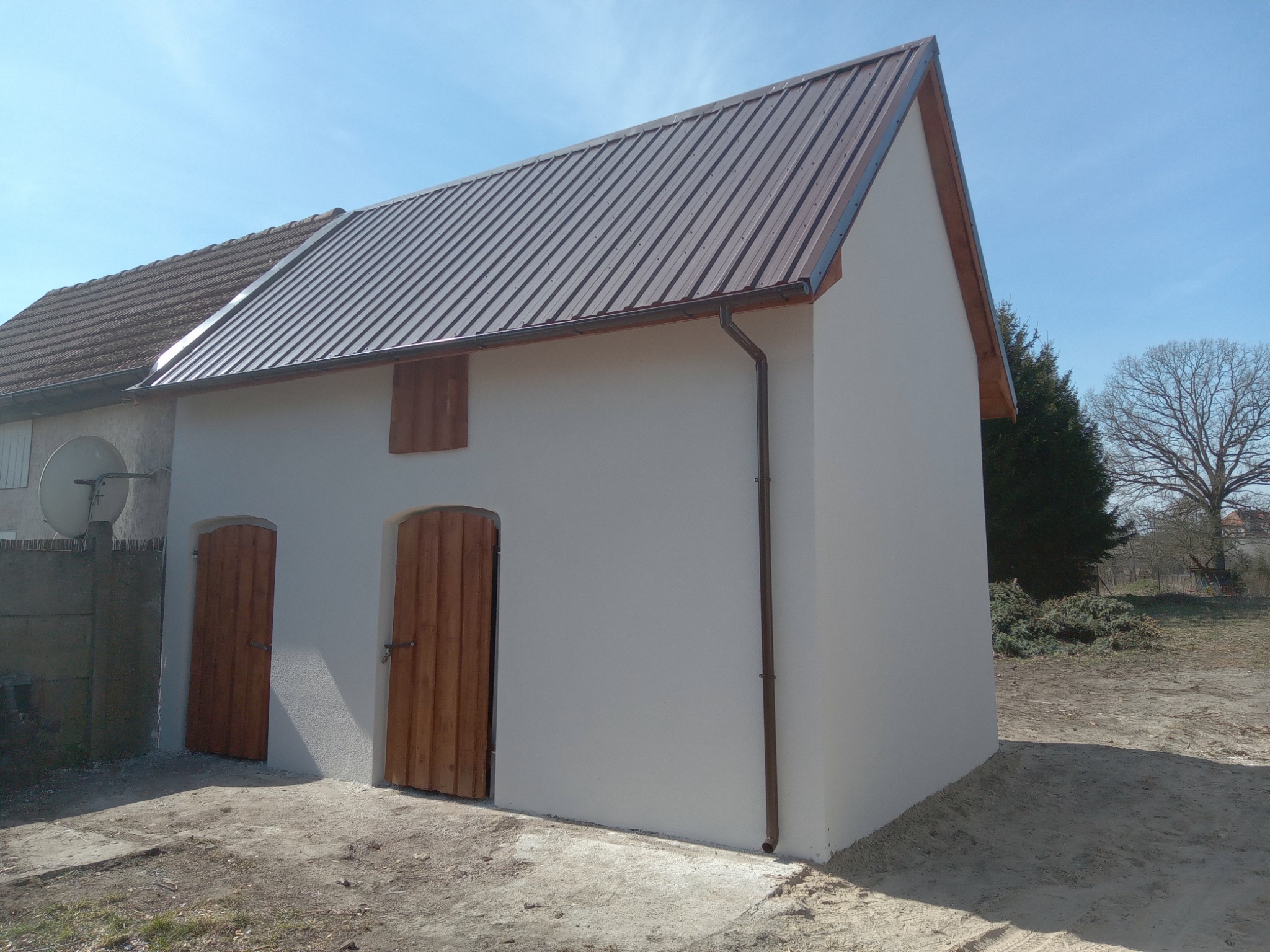 Świeżo otynkowany budynek gospodarczy z brązowym, metalowym dachem i drewnianymi drzwiami w rustykalnym stylu, widoczny fragment starego budynku z anteną satelitarną w tle.