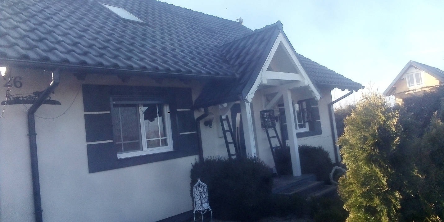 Dwukondygnacyjny dom z ciemną dachówką i jasną elewacją, widoczne okna z ciemnymi okiennicami, drabiny opierające się o ścianę przy wejściu, dekoracyjna klatka dla ptaków na zewnątrz.